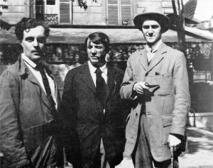 Modigliani, Picasso and André Salmon - LeFrancophile Three men in early 20th-century suits stand outside a Montparnasse building with shuttered windows. The man on the right wears a hat and holds a pipe, while the other two stand close together, all looking toward the camera with neutral expressions.