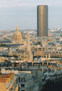 Tour montparnasse - LeFrancophile A view of Paris showing historic rooftops, a church with a tall spire in the center, the golden dome of Les Invalides to the left, and Montparnasse Tower rising in the background against a pale sky.