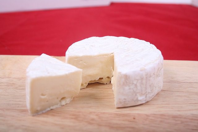 - LeFrancophile Une meule ronde de brie à pâte molle et à croûte fleurie repose sur une planche en bois. Une part est coupée, révélant l'intérieur crémeux.