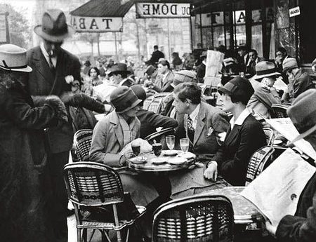 Black-and-white photo of a crowded Parisian café terrace in the 1920s. People in vintage clothing sit at round tables with drinks, talking and reading newspapers. A waiter serves a group, and wicker chairs fill the lively outdoor scene under awnings.